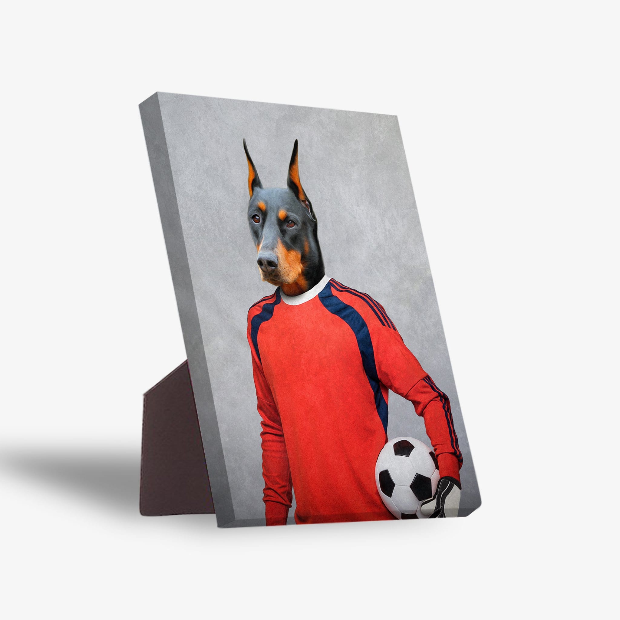 Doberman pet portrait on a standing canvas, dressed as a soccer goalie in a red jersey, holding a ball.